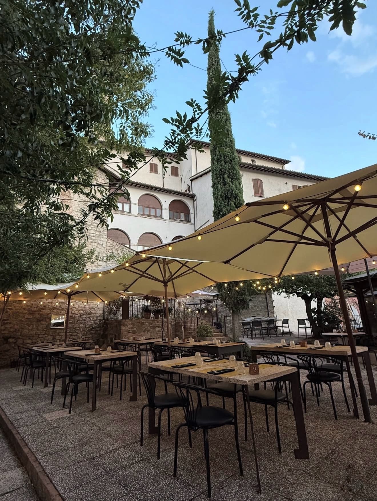 La corte la sera, tra gli ombrelloni e le luci, con il cipresso e il palazzo storico sullo sfondo